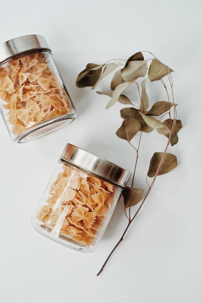 Minimalist flat lay of bowtie pasta in glass jars with dried leaves on a light background.